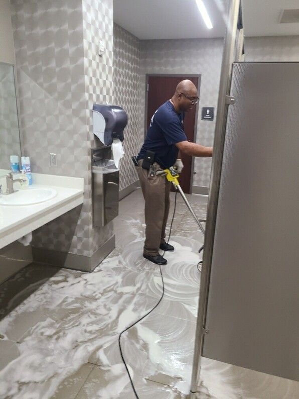 Worker cleaning a restroom floor with a pressure washer, beside sinks and stalls.