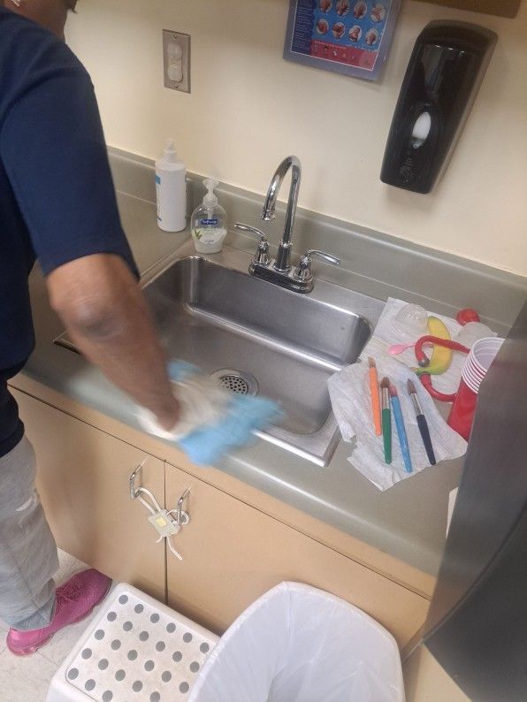 Person wiping a stainless steel sink in a clinic or kitchen, with cleaning supplies nearby.