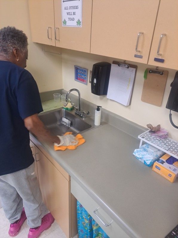 Person wiping a kitchen counter and sink with an orange cloth in a beige break room.