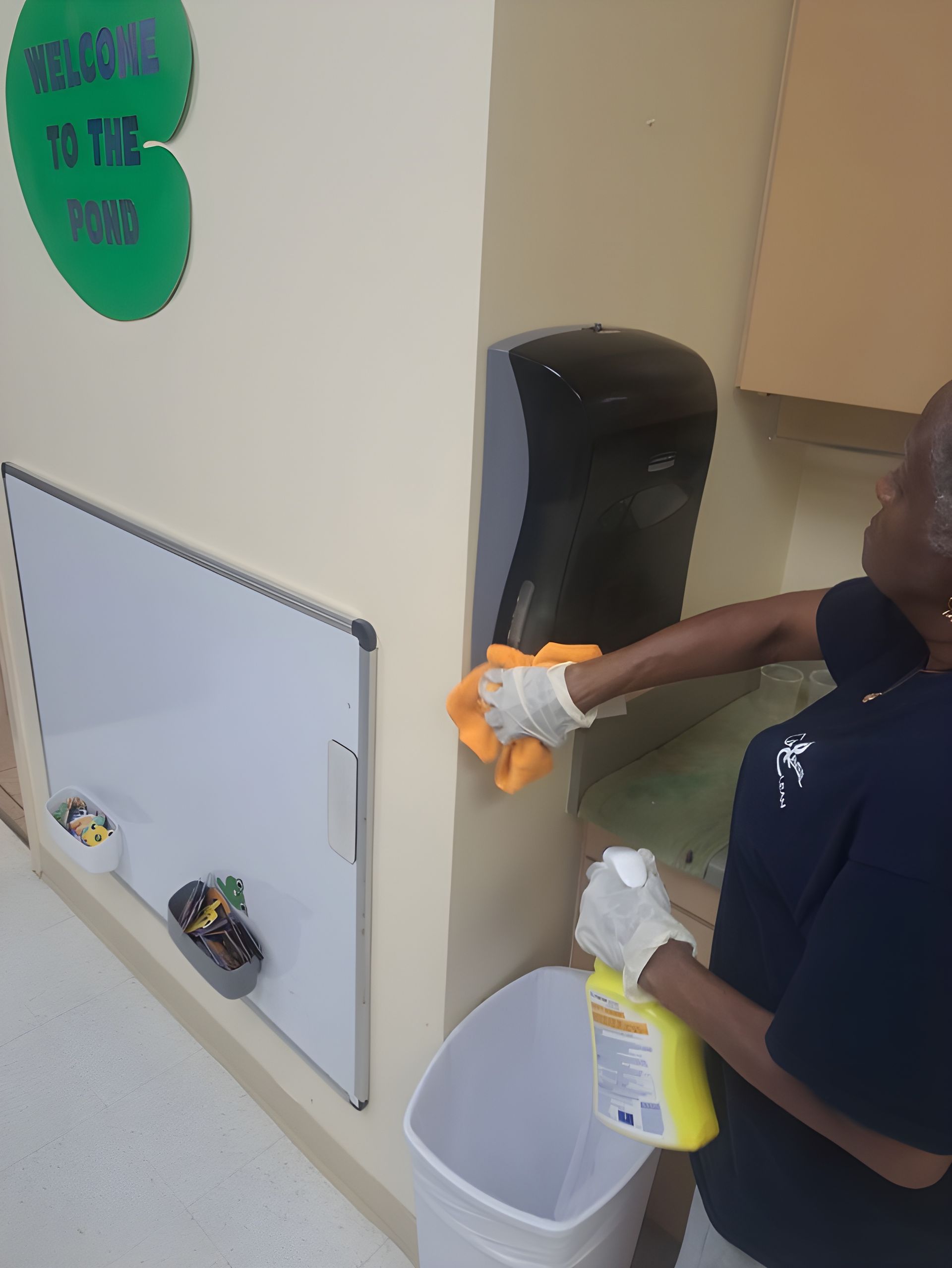 Child cleaning a wall-mounted dispenser with spray bottle and cloth in a classroom corner