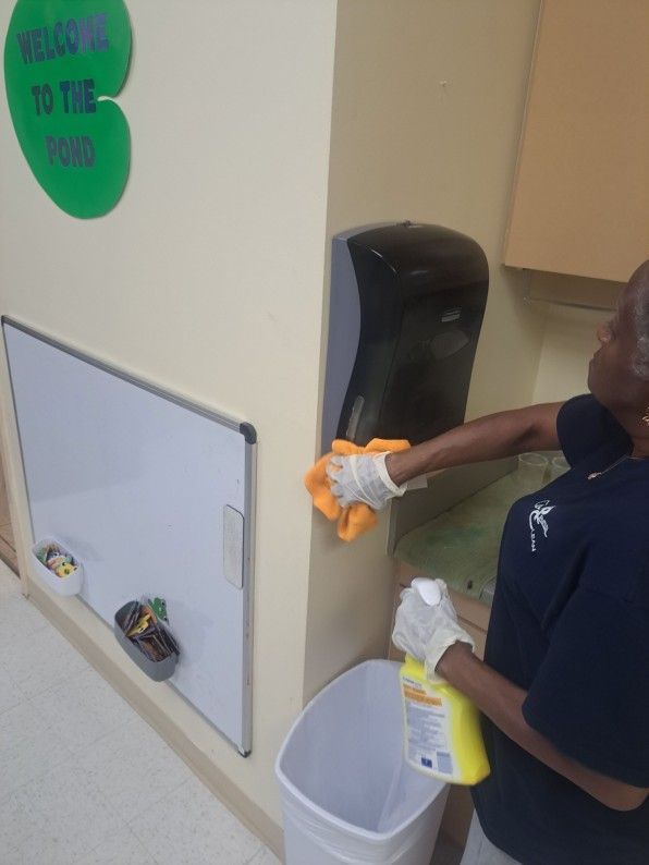 Person cleaning a restroom wall with a spray bottle and cloth beside a trash bin