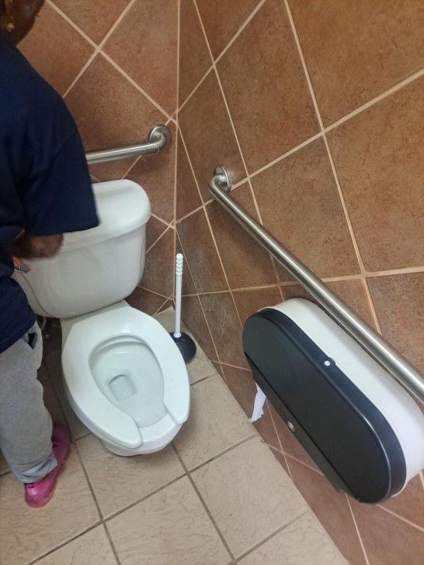 Public restroom stall with open toilet, grab bar, and wall-mounted dispenser; a person stands beside it.