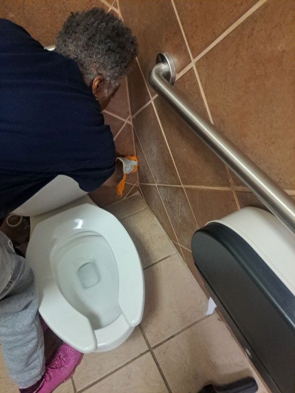 Person cleaning a restroom floor beside a toilet and grab bar.