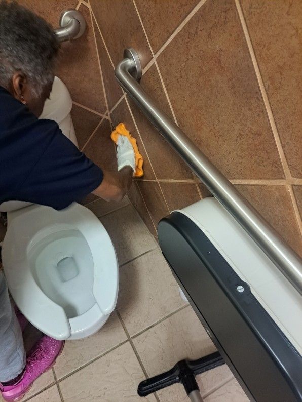 Person cleaning a restroom floor beside a toilet and grab bar with a cloth and spray bottle