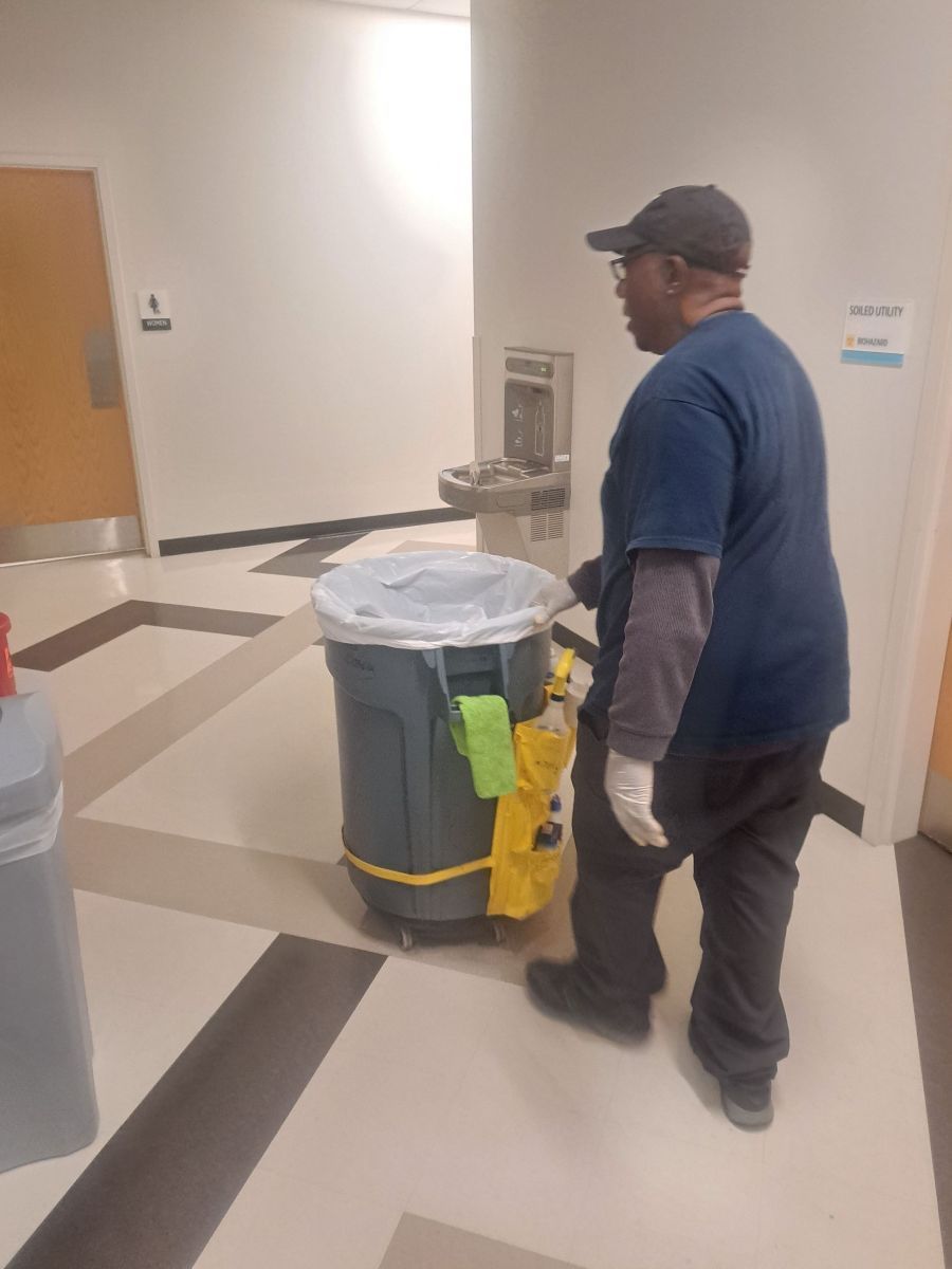 Custodian in blue shirt pushing a trash bin through a hallway with a checkered floor.
