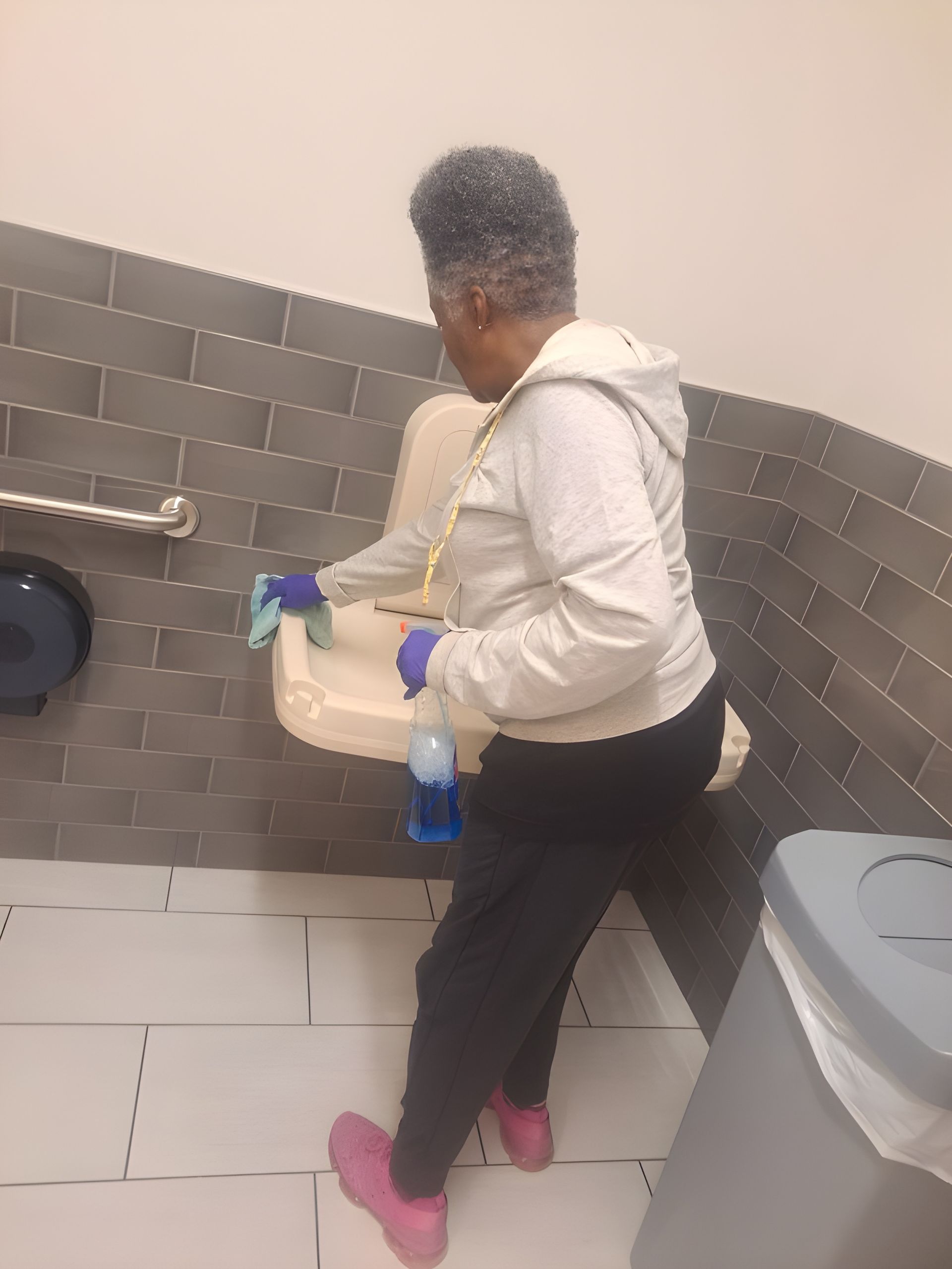 Person cleaning a bathroom sink, wearing gloves, white hoodie, black pants, and pink shoes.