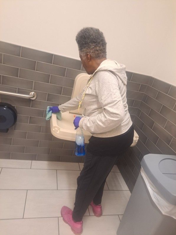 Person cleaning a restroom sink with a spray bottle and cloth, wearing a hoodie, gloves, and pink shoes.