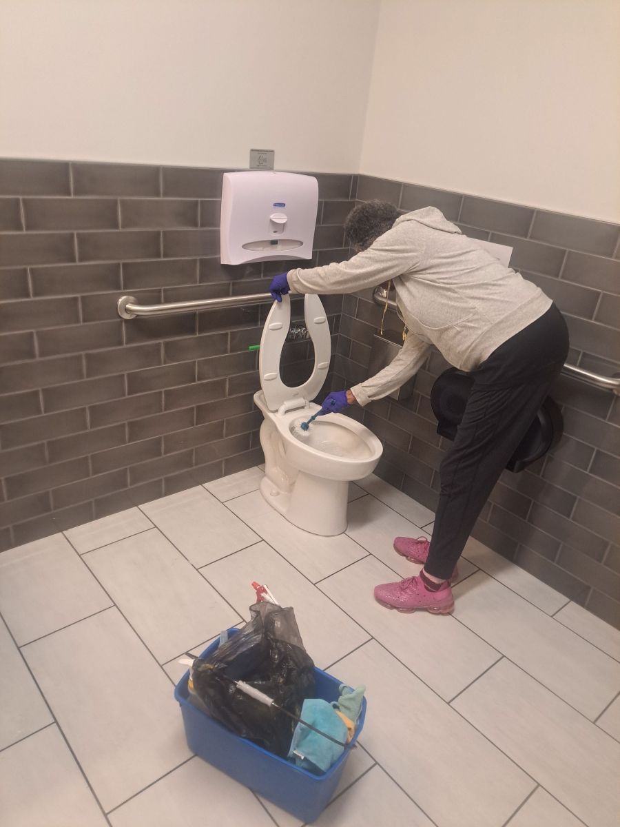 Person cleaning a toilet in a public restroom, with a blue cleaning caddy on the floor.