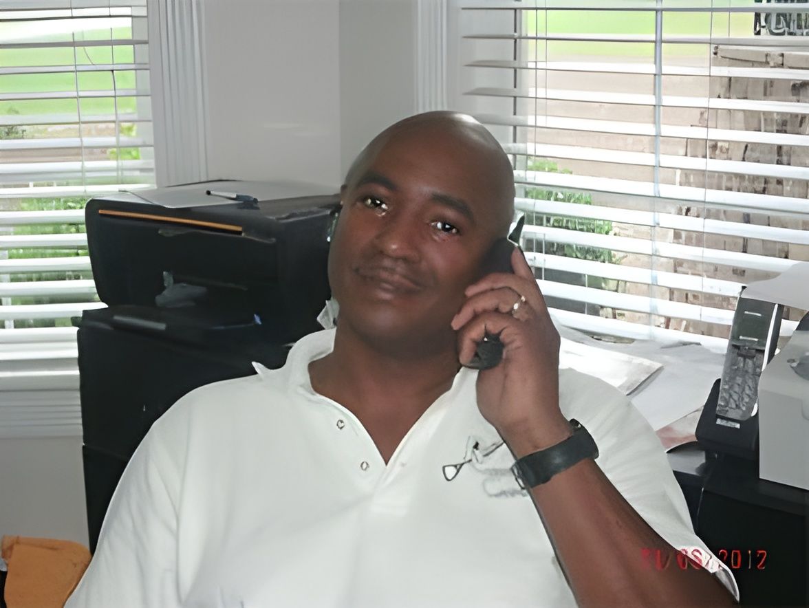 Man in a white polo shirt talking on a phone at a desk in a bright office