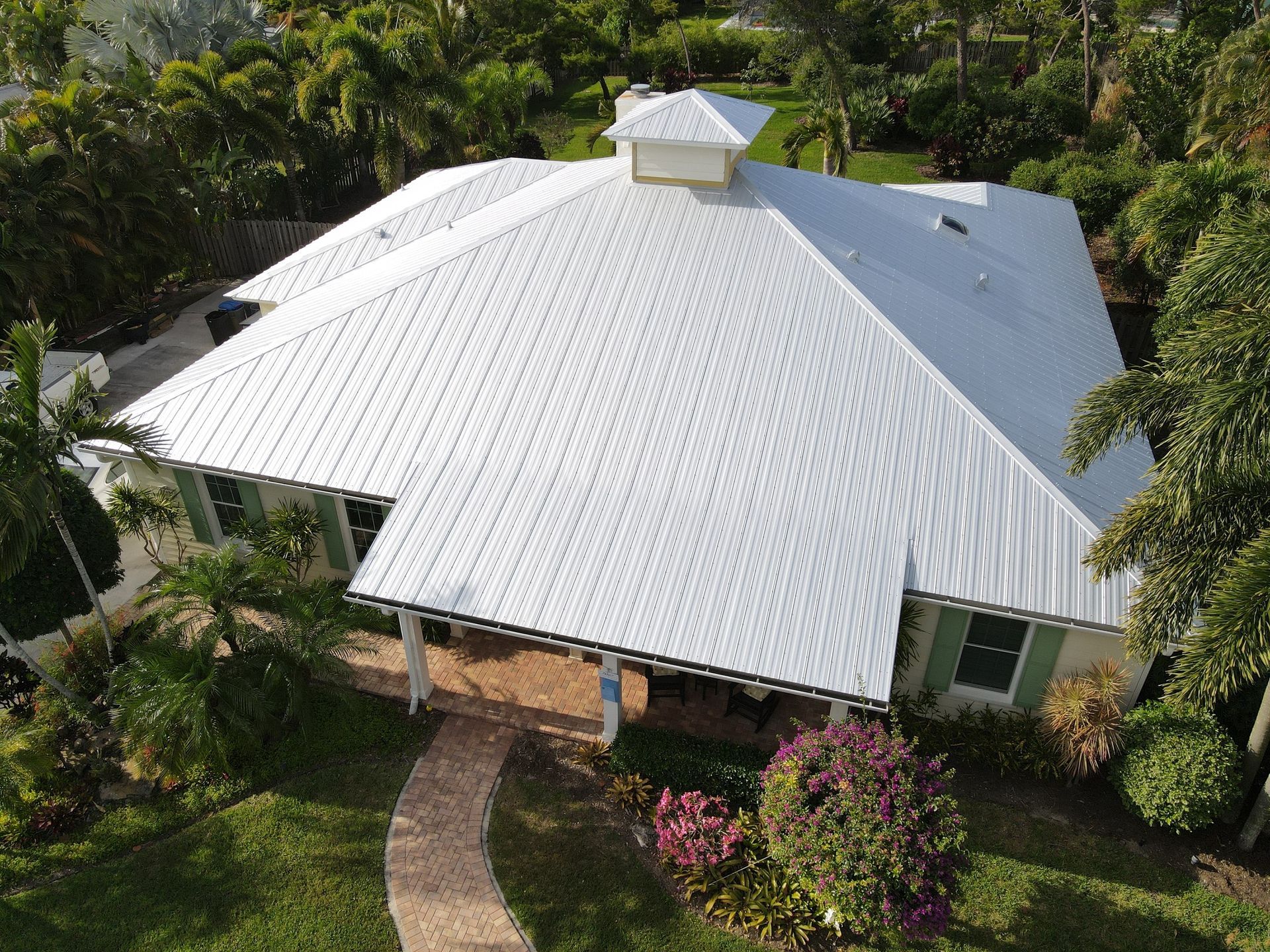 Aerial view of a white metal-roofed house surrounded by lush green trees and a curved walkway.
