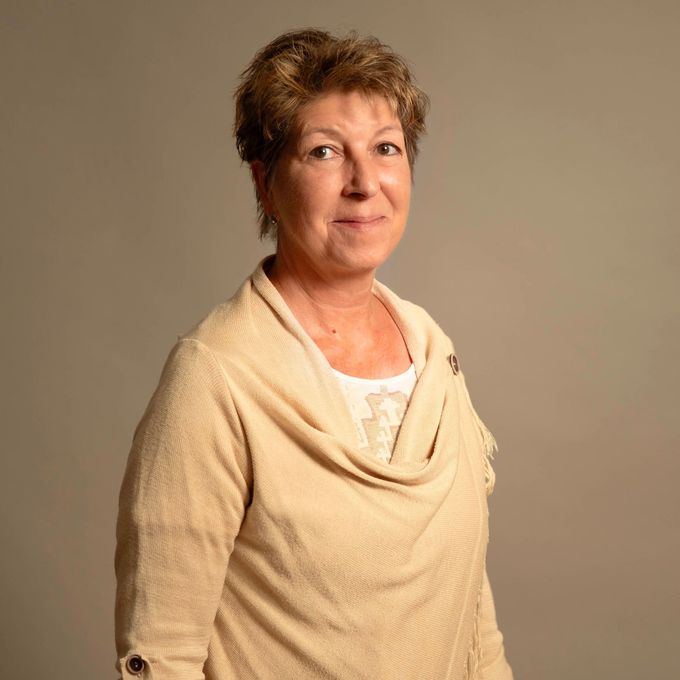 Frau in einem beigen Pullover posiert vor einem schlichten grauen Hintergrund.