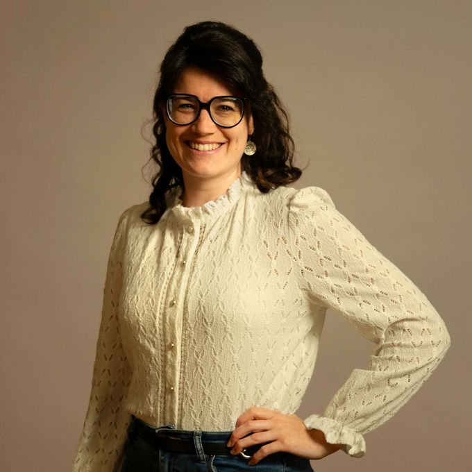 Lächelnde Frau mit Brille, bekleidet mit einer cremefarbenen Bluse und dunklen Jeans, vor beigem Hintergrund.
