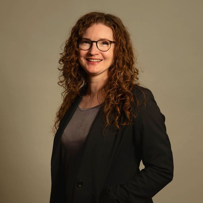Lächelnde Frau mit lockigem Haar und Brille in einem schwarzen Blazer vor schlichtem beigefarbenem Hintergrund