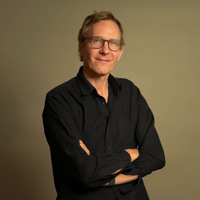 Studioaufnahme eines Mannes in schwarzem Hemd und Brille, die Arme verschränkt, vor beigem Hintergrund