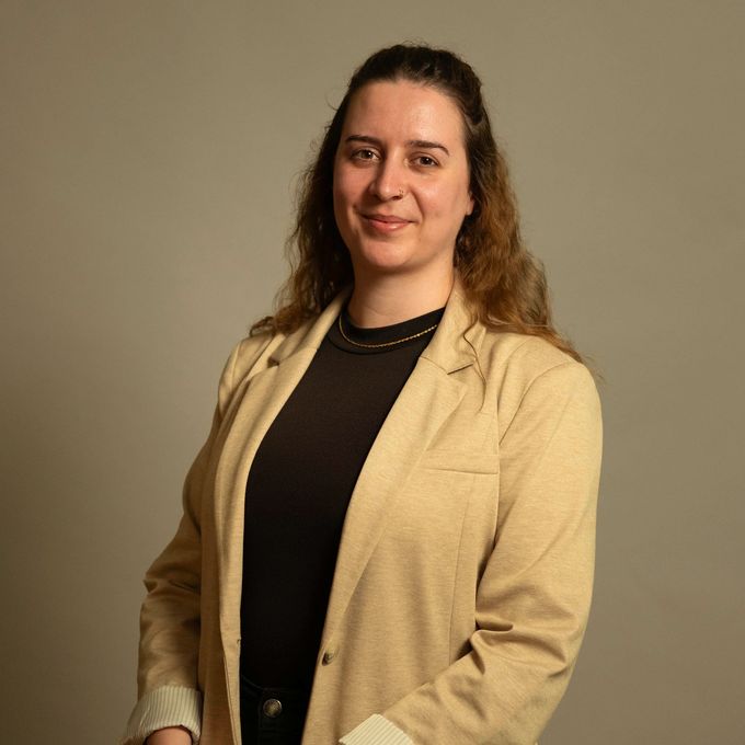 Eine Frau steht vor einem neutralen Hintergrund, trägt einen beigen Blazer über einem schwarzen Oberteil und lächelt sanft.