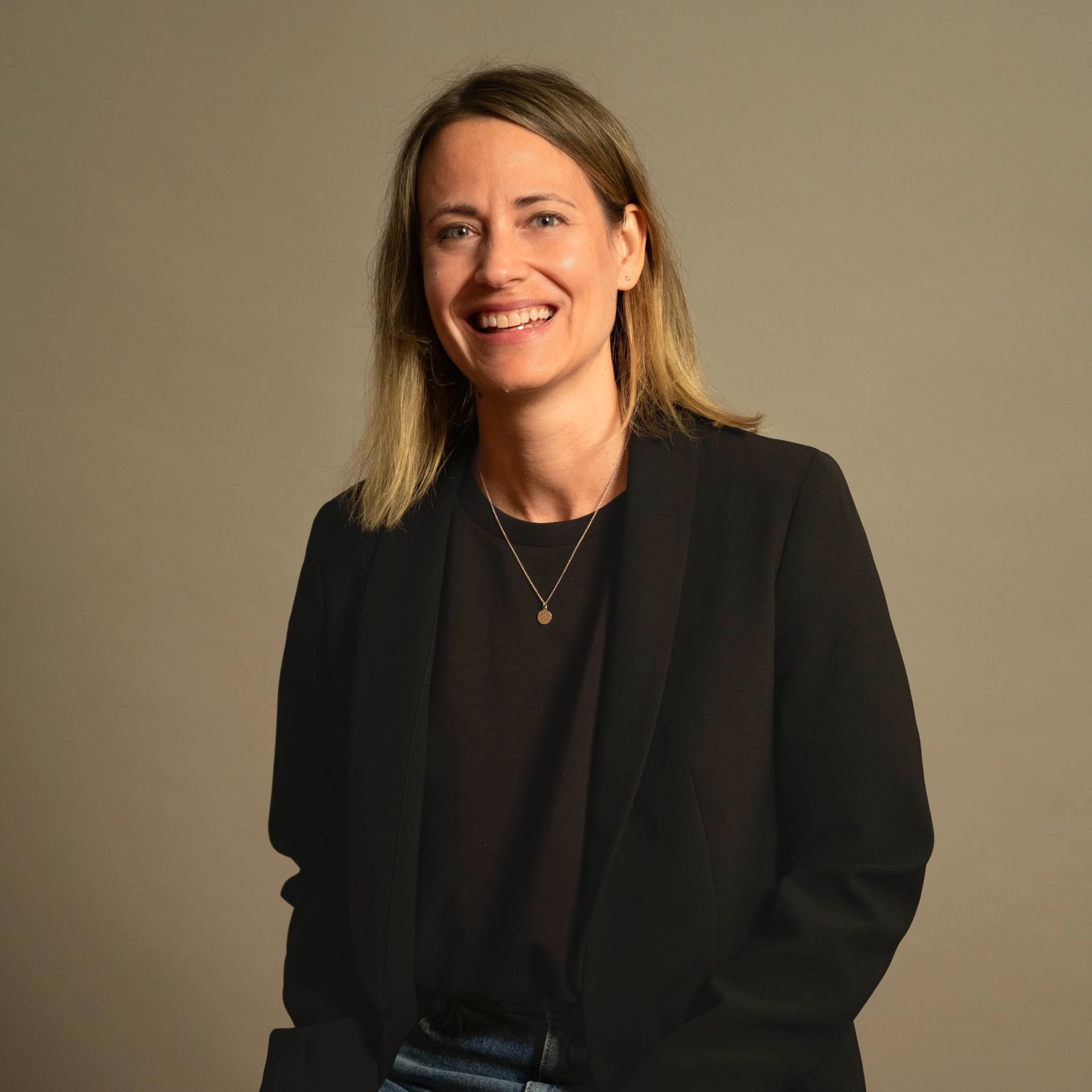 Frau in einem schwarzen Blazer lächelt vor einem schlichten beigen Hintergrund