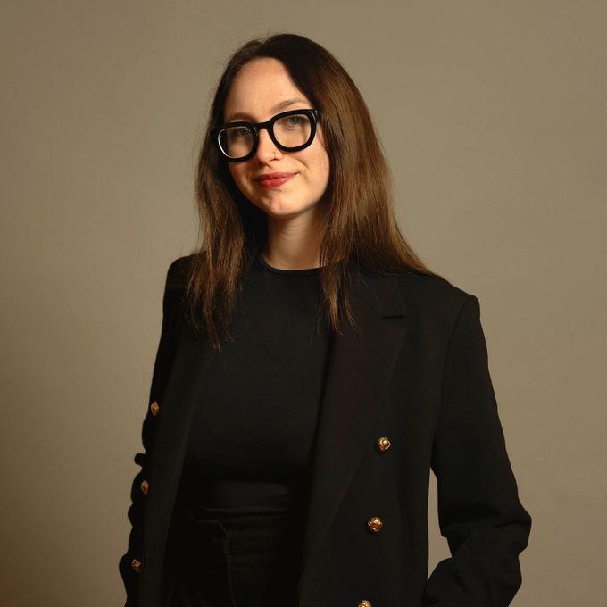 Frau in schwarzem Blazer und Brille, vor einem schlichten beigen Hintergrund.