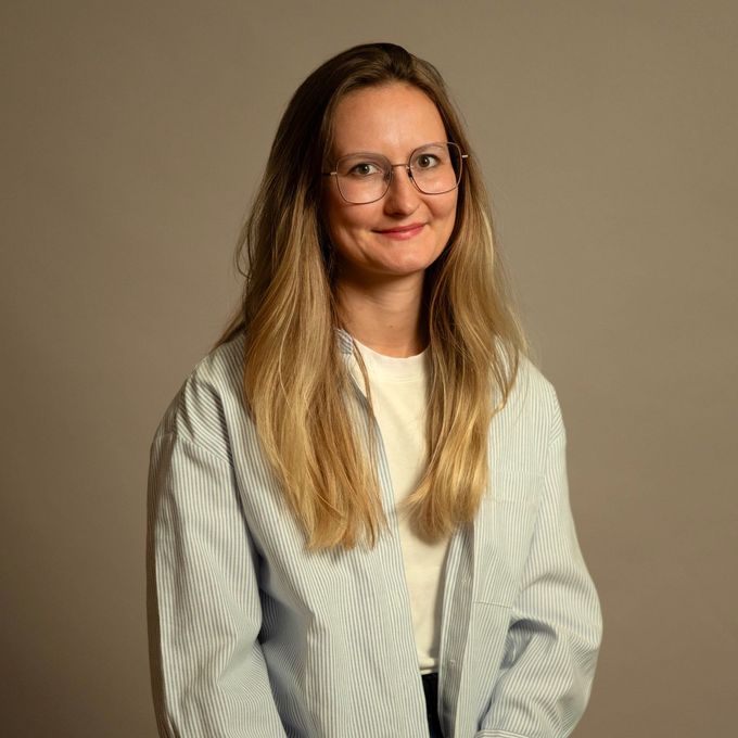 Frau mit langen blonden Haaren und Brille, die ein hell gestreiftes Hemd vor einem einfarbigen braunen Hintergrund trägt