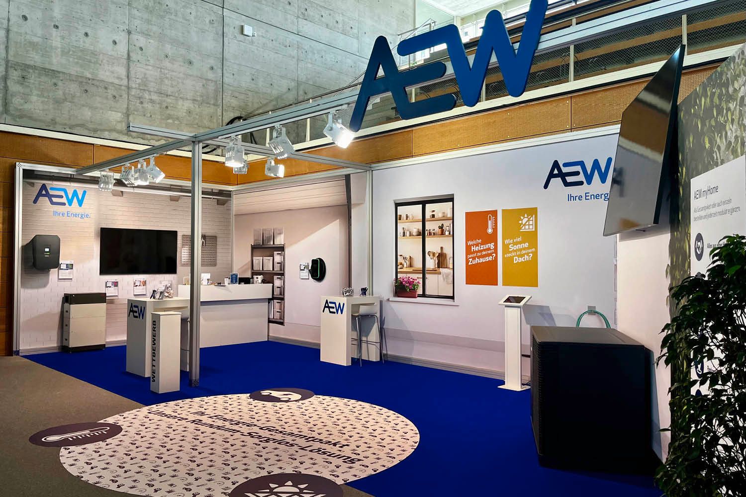 AEW-Messestand mit blauem Teppich, weißen Displays, großem Logo und Hängeschild