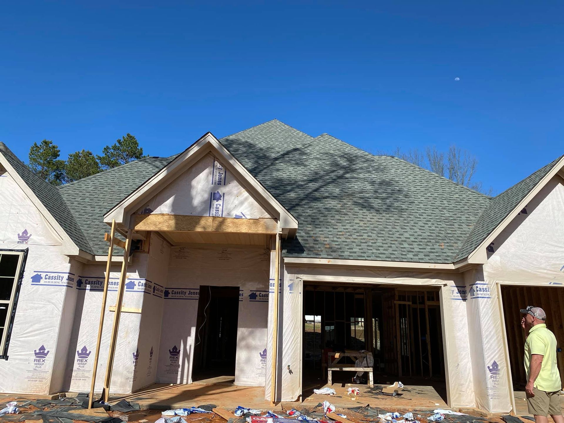 House under construction with a gray roof and blue sky. A worker stands on the right.