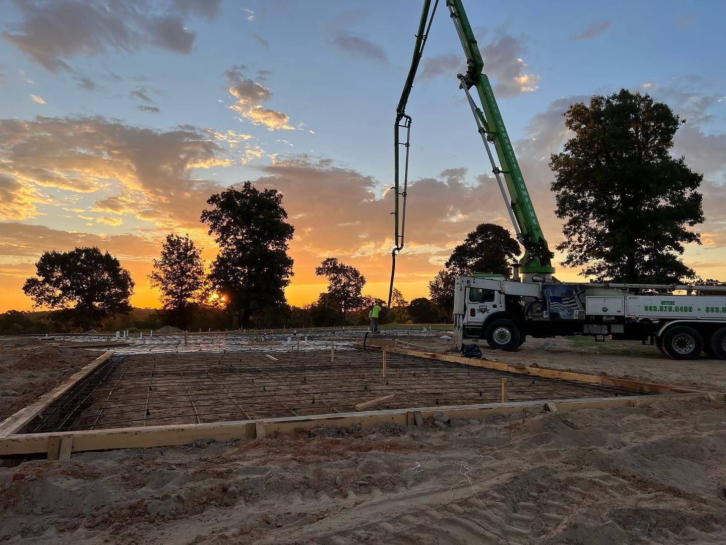 Construction site at sunset with concrete being poured into foundation. A truck with a crane is in use.