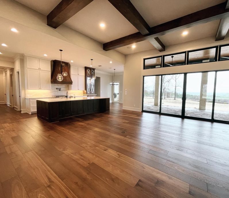 Open-concept kitchen and living area with wood floors, exposed beams, and large windows overlooking a landscape.