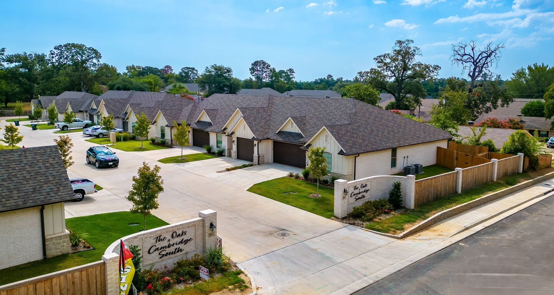Row of townhomes, tan siding, brown roofs, gated entrance, green lawns, blue sky.
