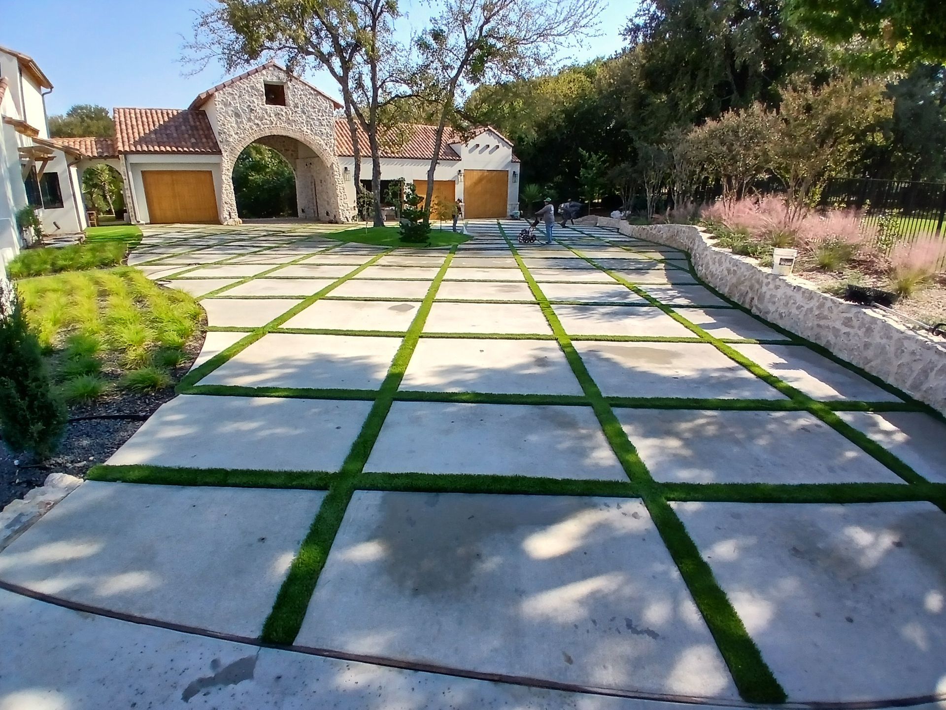 Sunny courtyard with stone pavers and grass strips leading to a house with a brick archway.