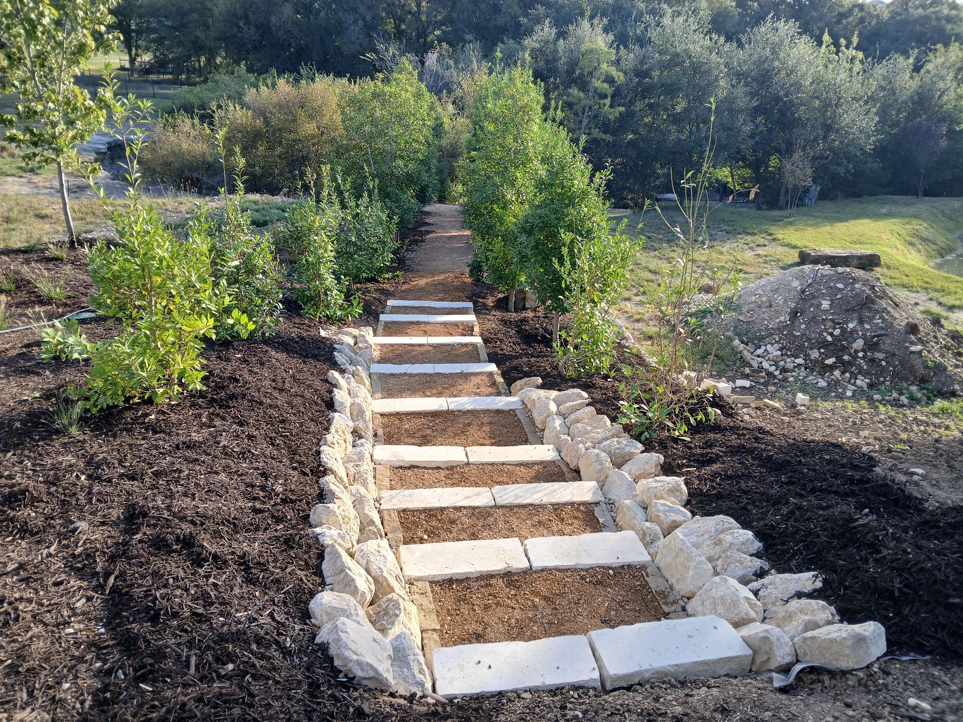 Stone garden steps bordered by rocks, leading up a dirt path through green shrubs and trees.