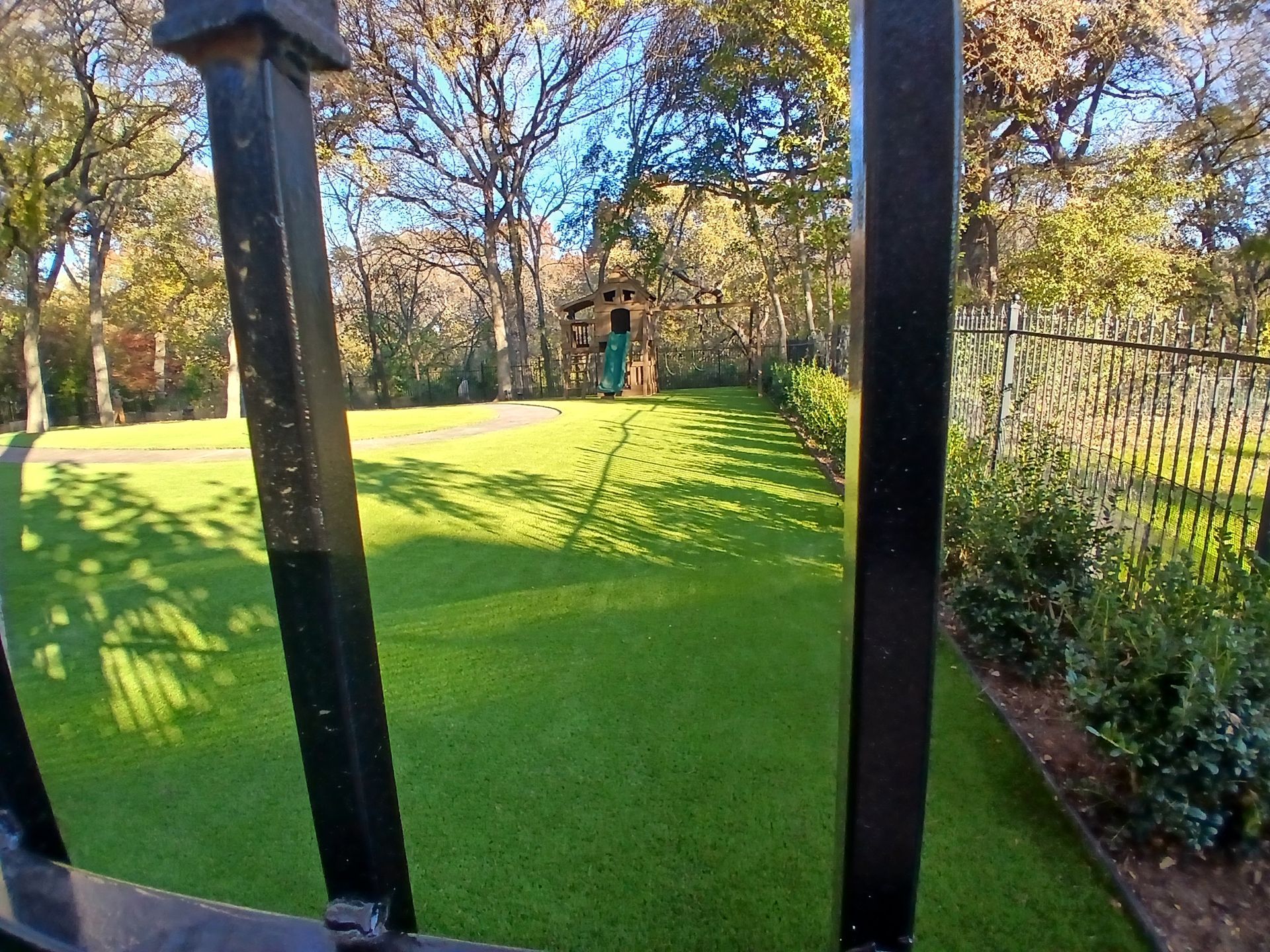Backyard with green lawn, black fence posts, and a child in the distance near trees and autumn leaves