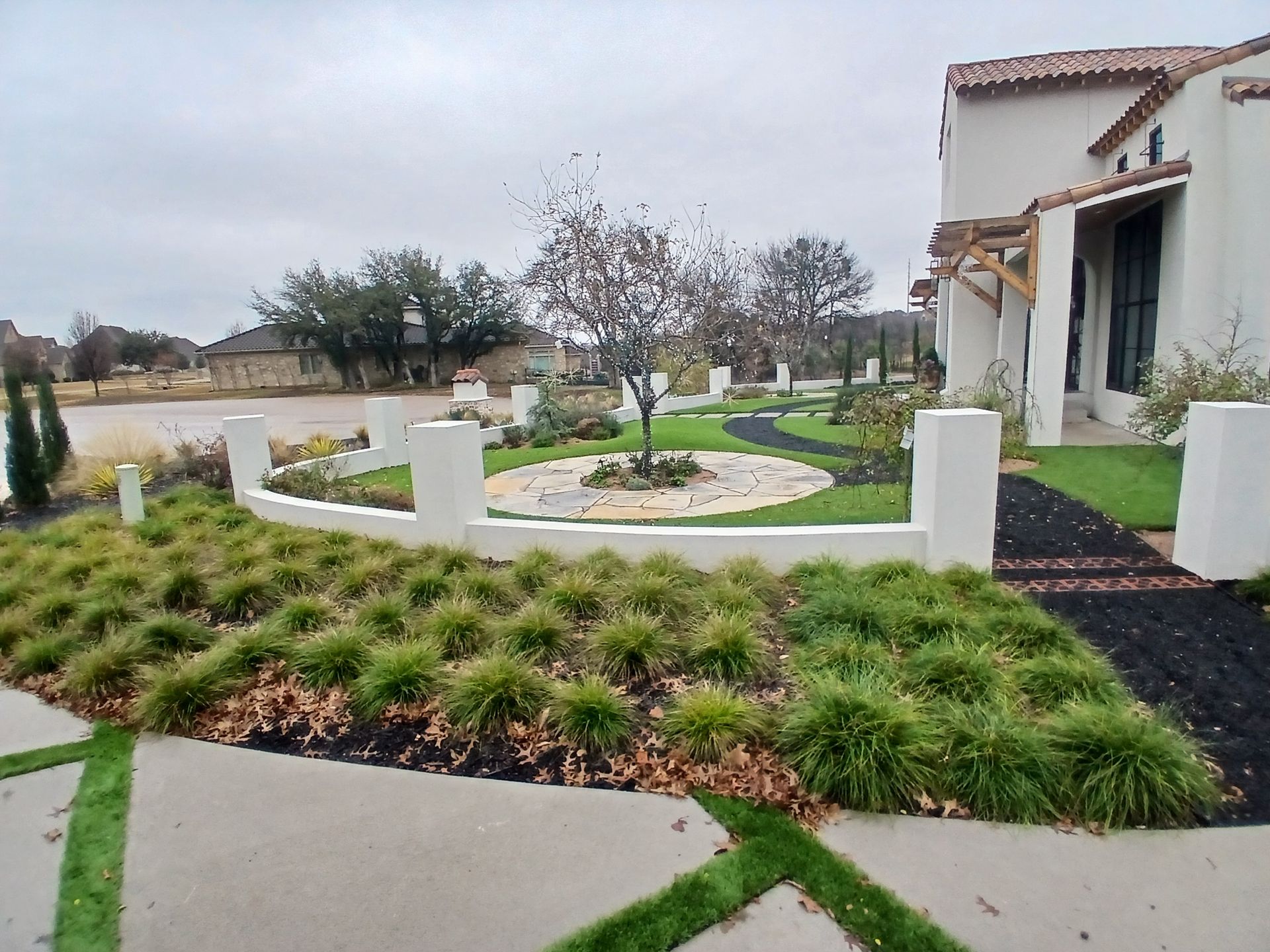 Front yard landscaping with white fence, green shrubs, curved walkway, and a house on an overcast day