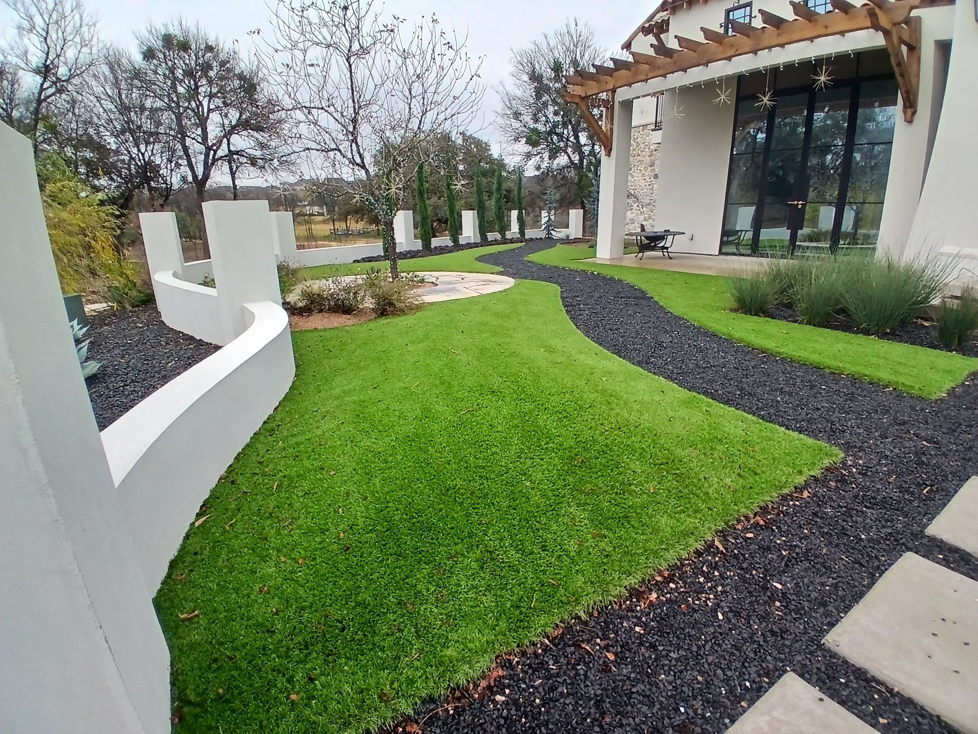 Curved black gravel path through a green lawn beside a modern house with white posts and glass doors