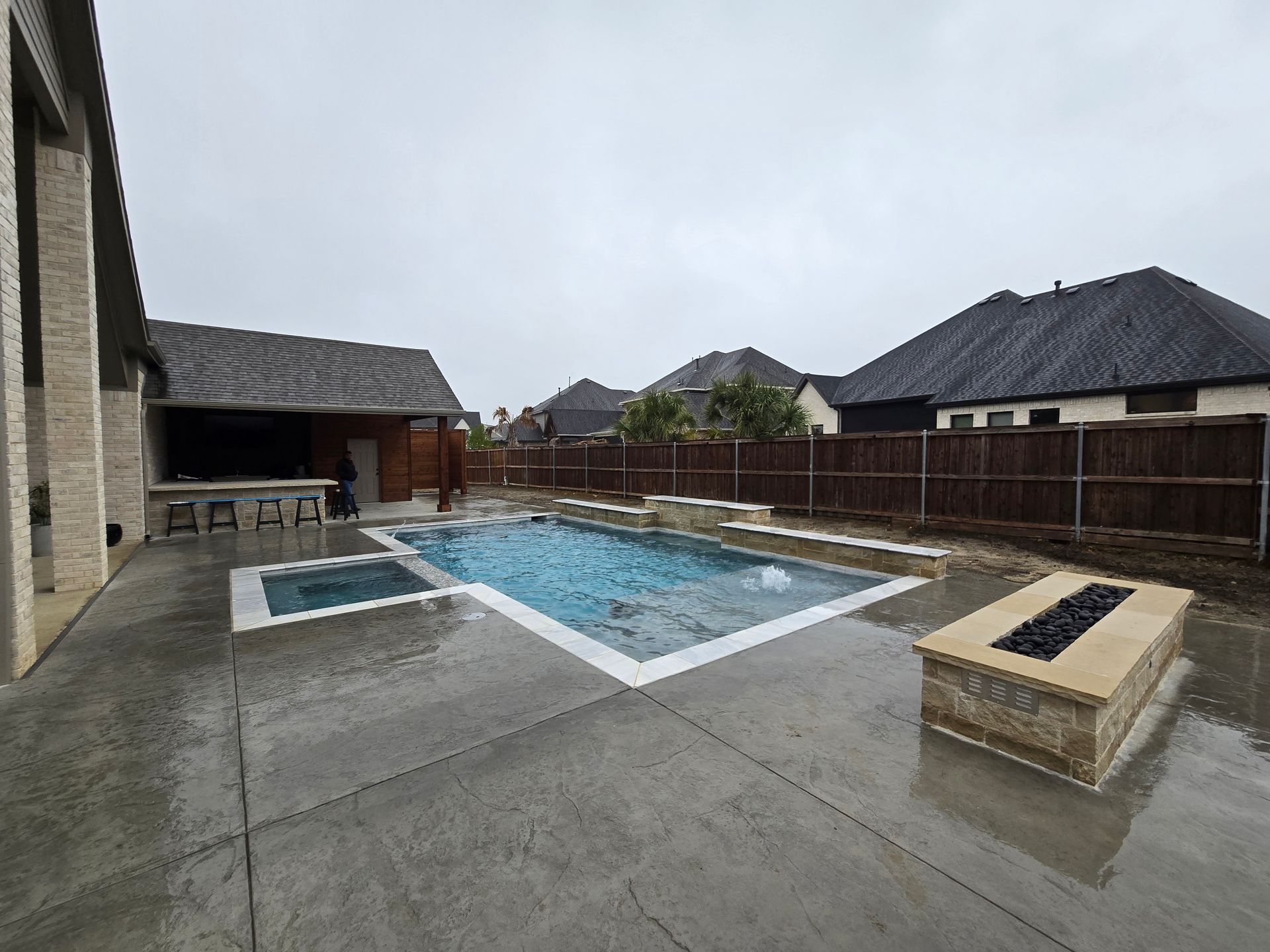 Backyard pool with a wet concrete deck, fenced perimeter, and overcast sky