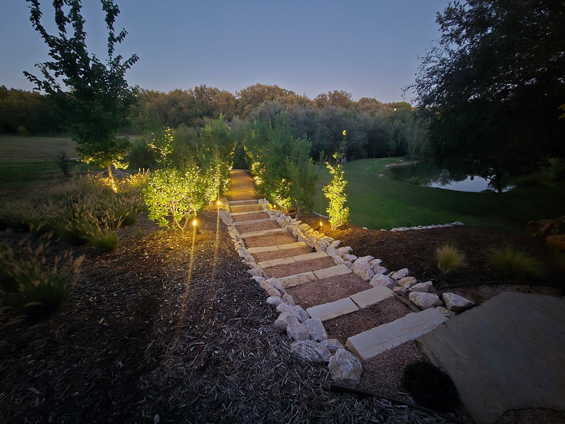 Stone steps lit by warm lights lead down a garden path at dusk, surrounded by grass and trees.