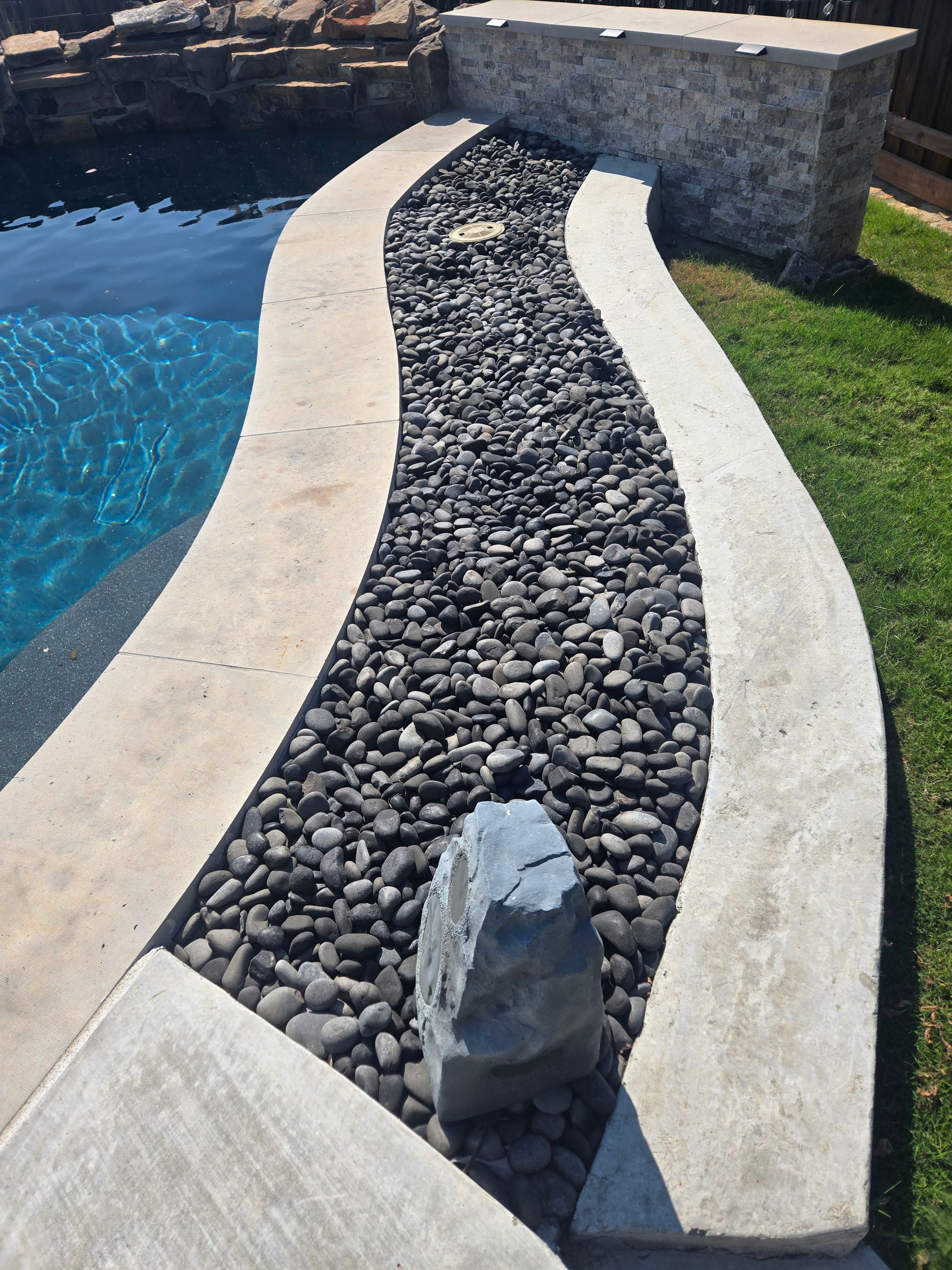 Curved stone walkway lined with black pebbles beside a blue pool and stone wall