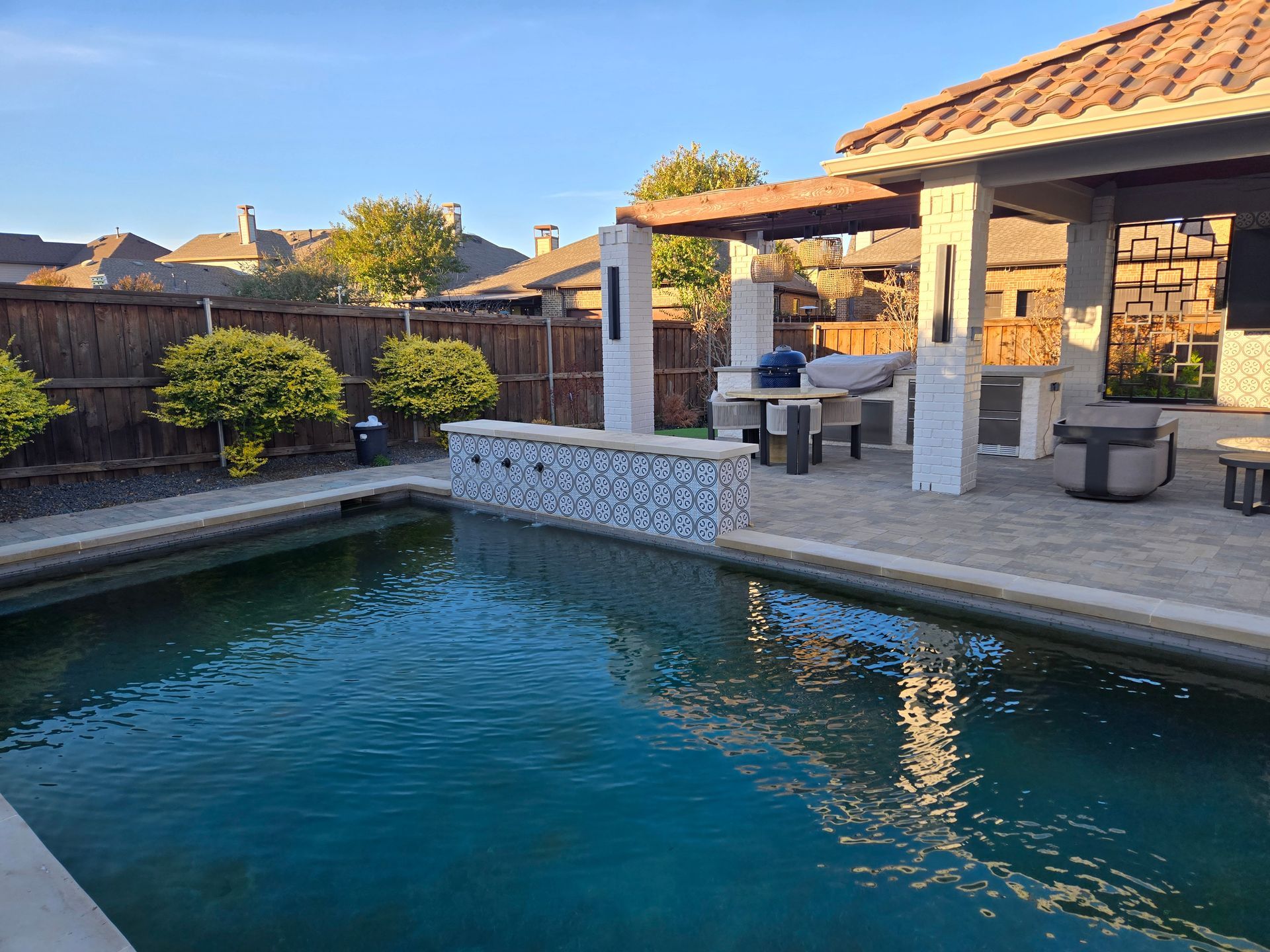 Backyard swimming pool beside a covered patio with stone seating and evening sunlight
