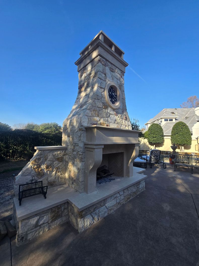 Stone outdoor fireplace and chimney under a bright blue sky in a courtyard with benches and trees
