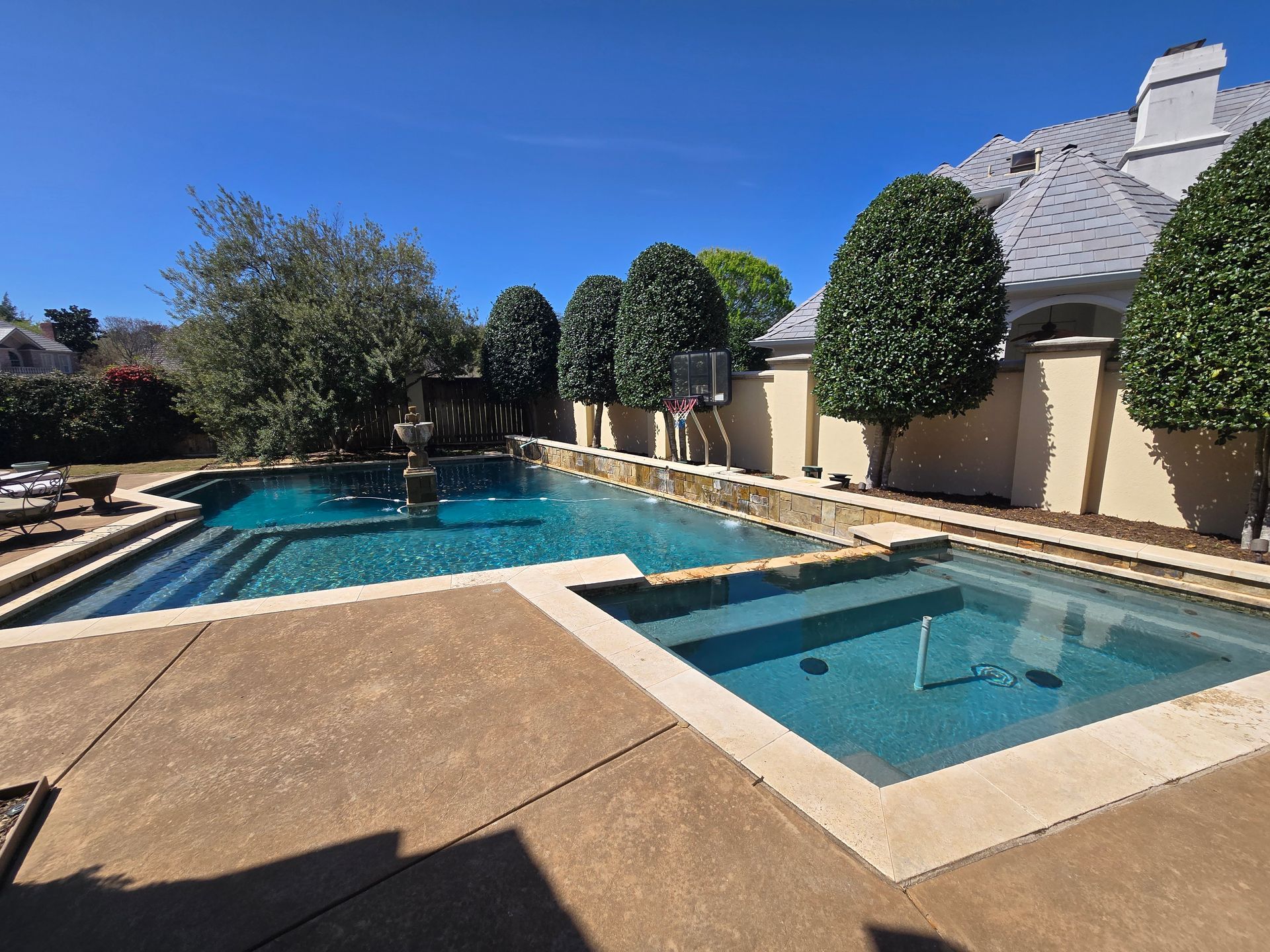 Backyard pool and hot tub beside a tan patio, trimmed trees, and a house under a clear blue sky