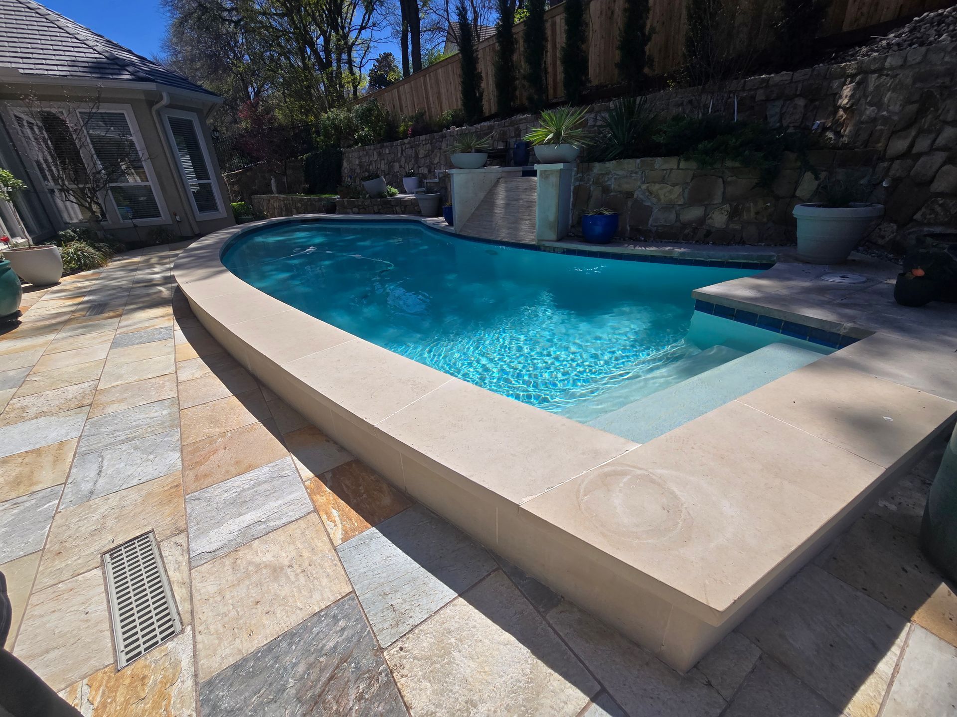 Backyard swimming pool with turquoise water, tan stone deck, and lounge chairs beside a garden wall