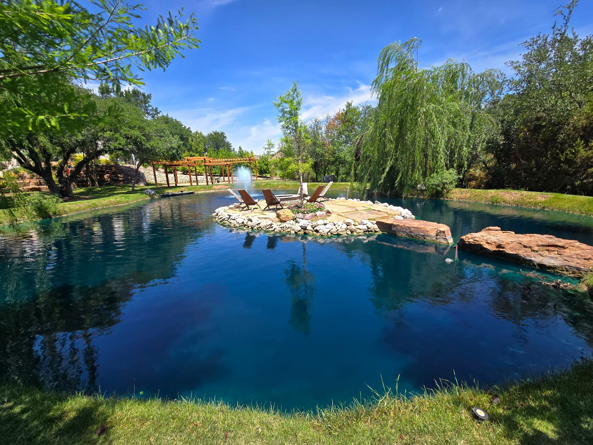 A blue lagoon pool with a small rock island, surrounded by trees under a clear sky.