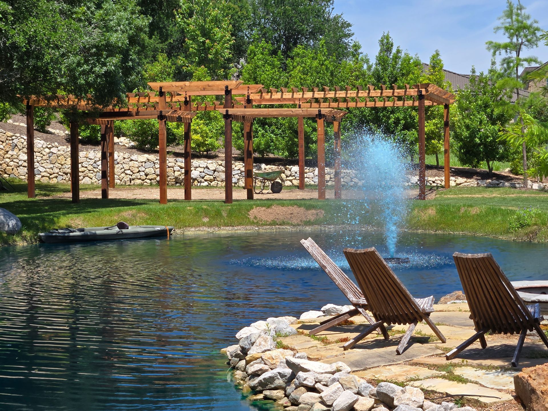 Lake with fountain, wooden pergola, and two lounge chairs on a stone patio beside the water
