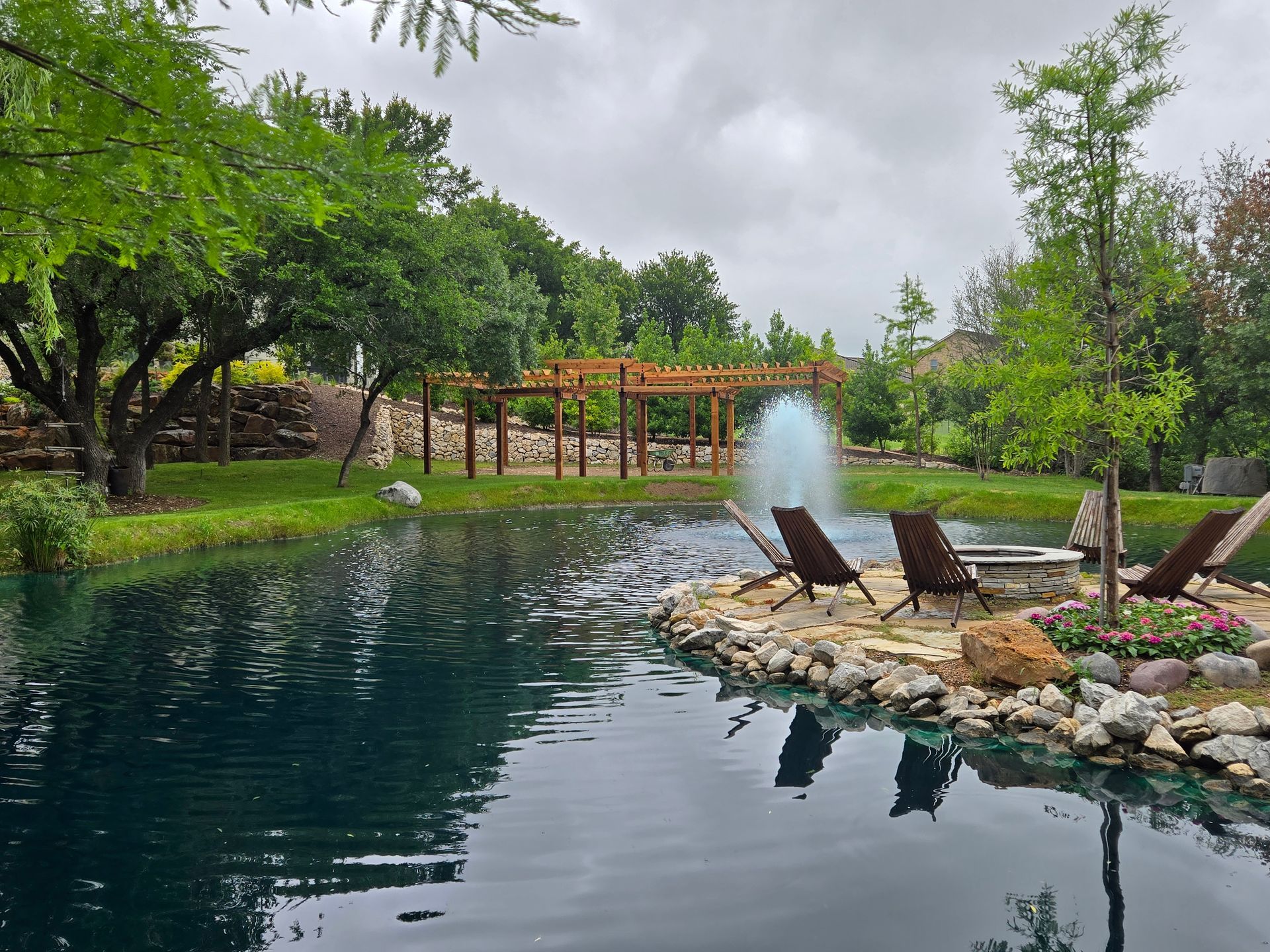 Serene garden pond with lounge chairs, a fountain, lush greenery, and a wooden pergola under cloudy skies