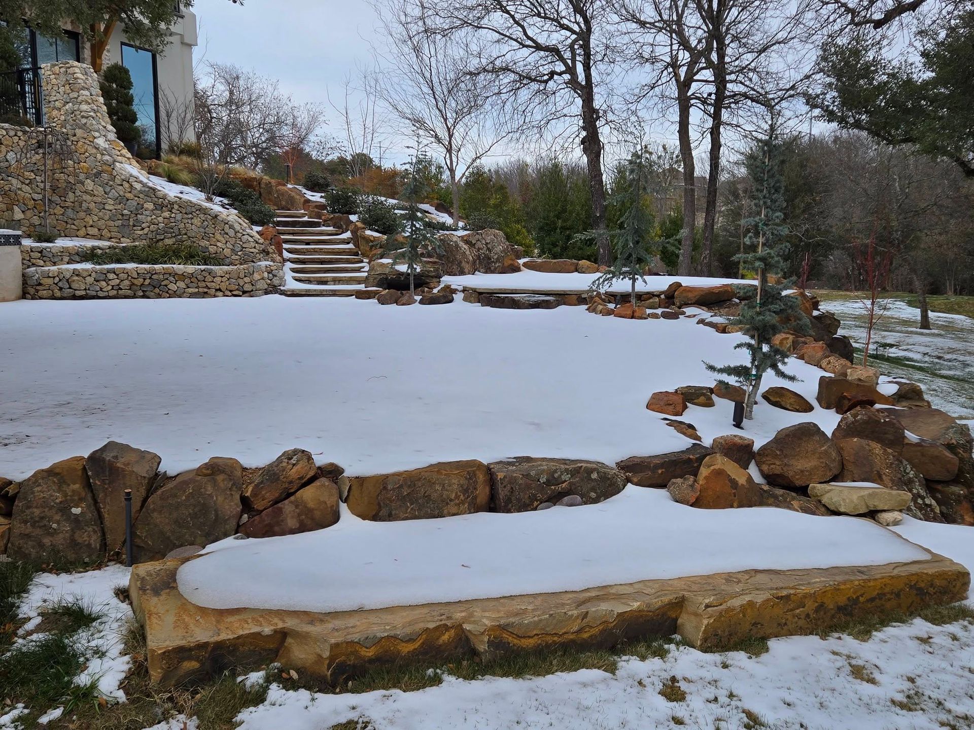 Snow-covered garden with stone steps, low retaining walls, and bare trees in winter.