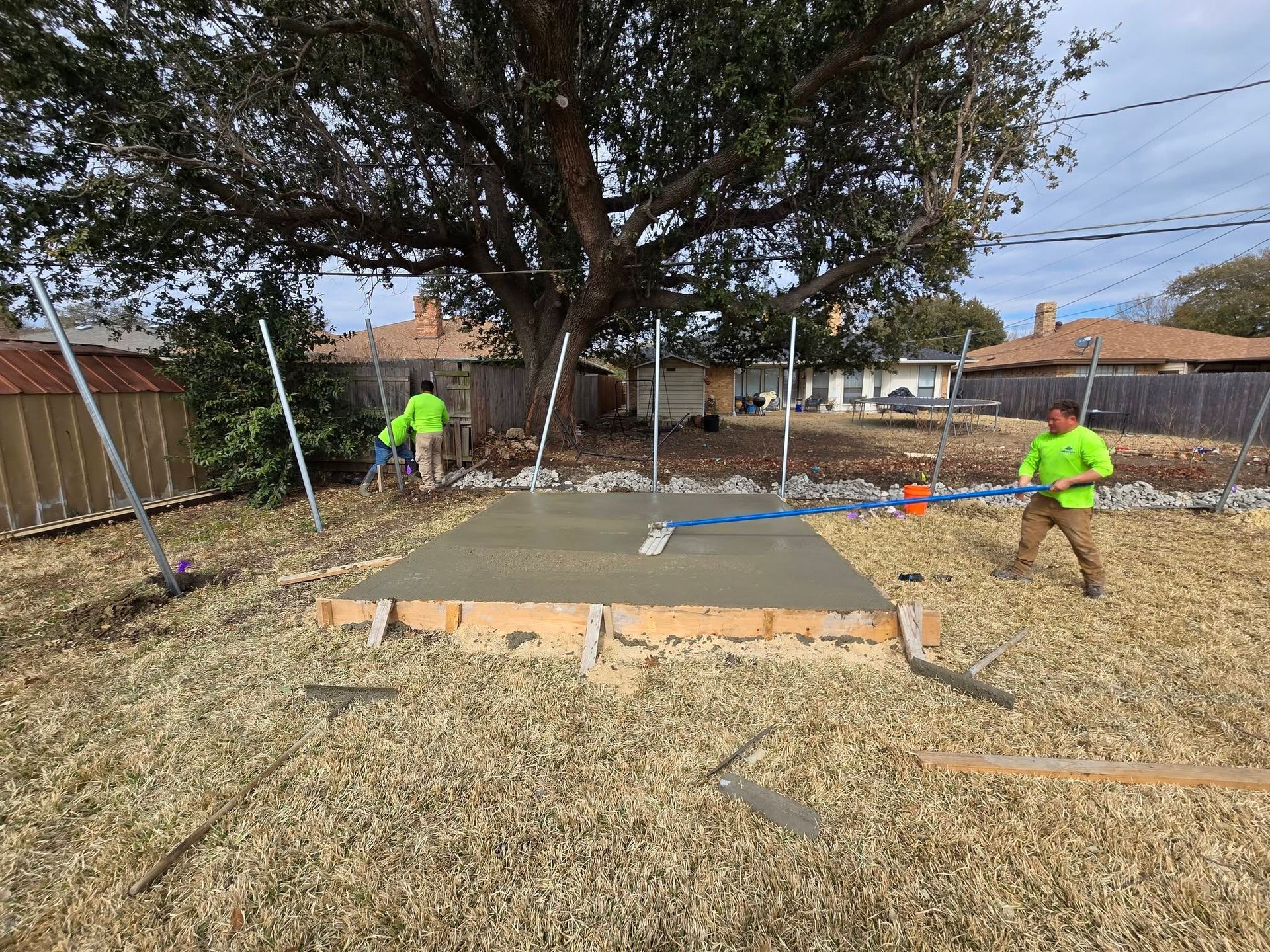 Workers pour concrete for a backyard patio under construction, with orange barriers and a tree overhead