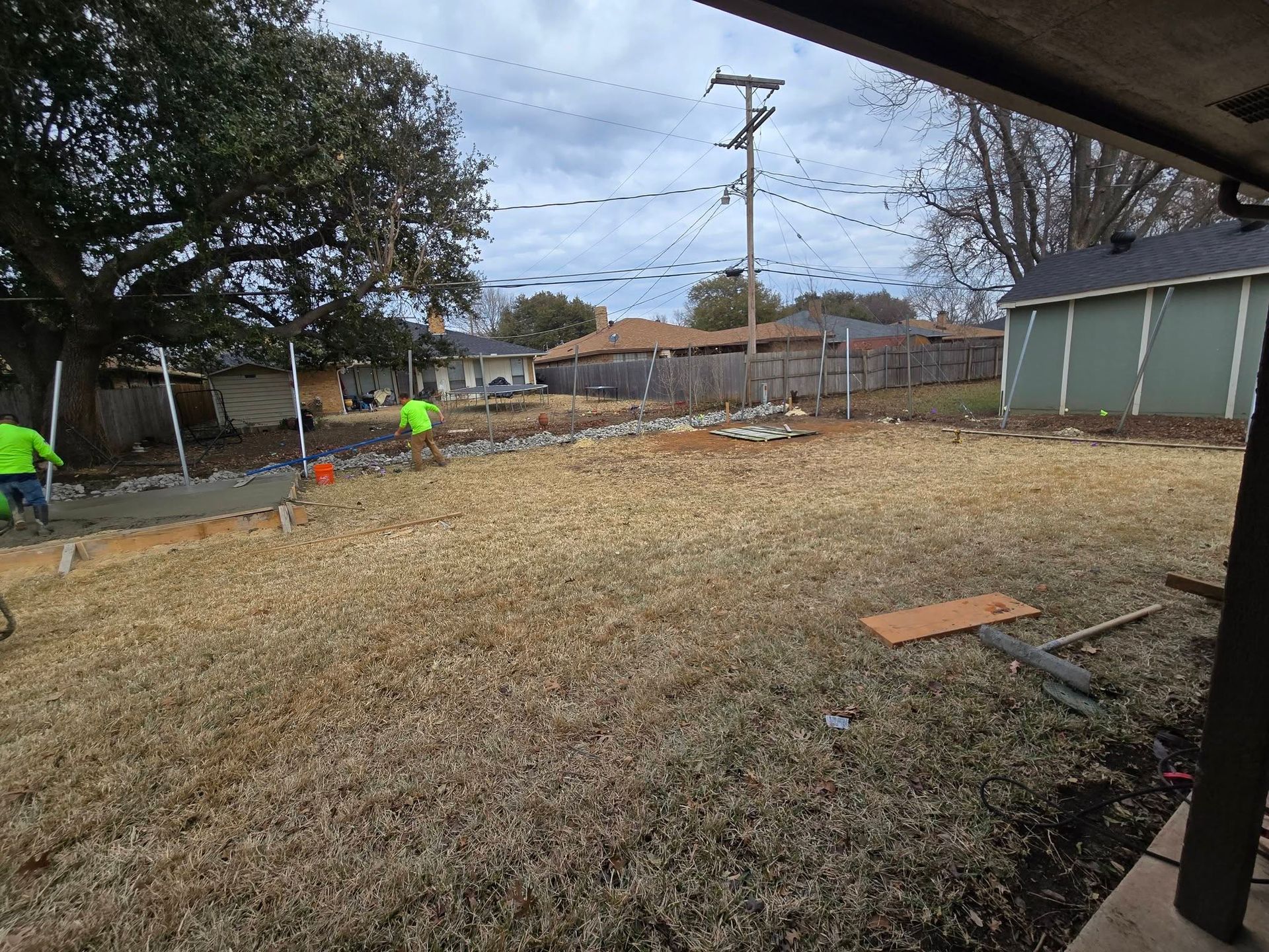 Backyard with workers installing a fence along the left side, surrounded by trees and sheds.
