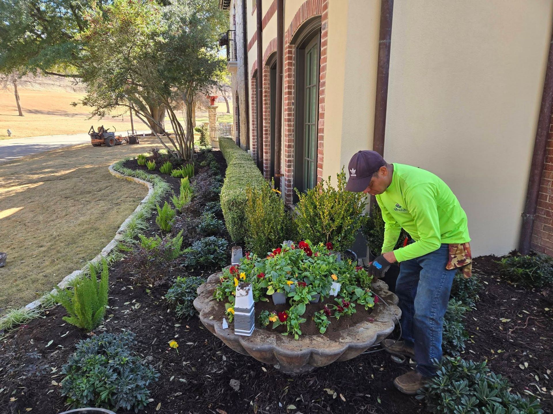 Person in a neon green shirt tending a flower bed beside a building.