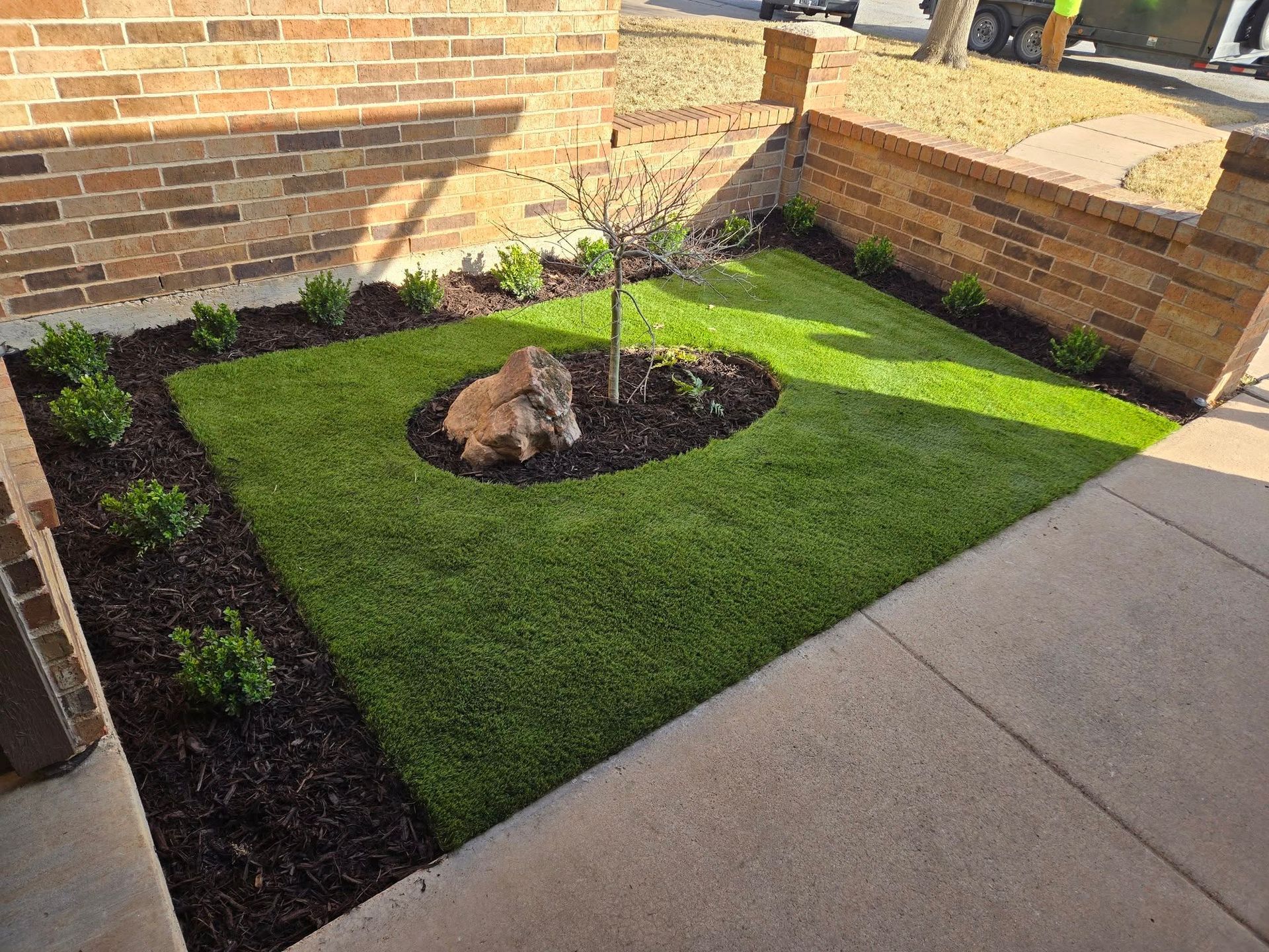 Small landscaped courtyard with fresh green turf, mulch beds, a young tree, and a rock on the grass.