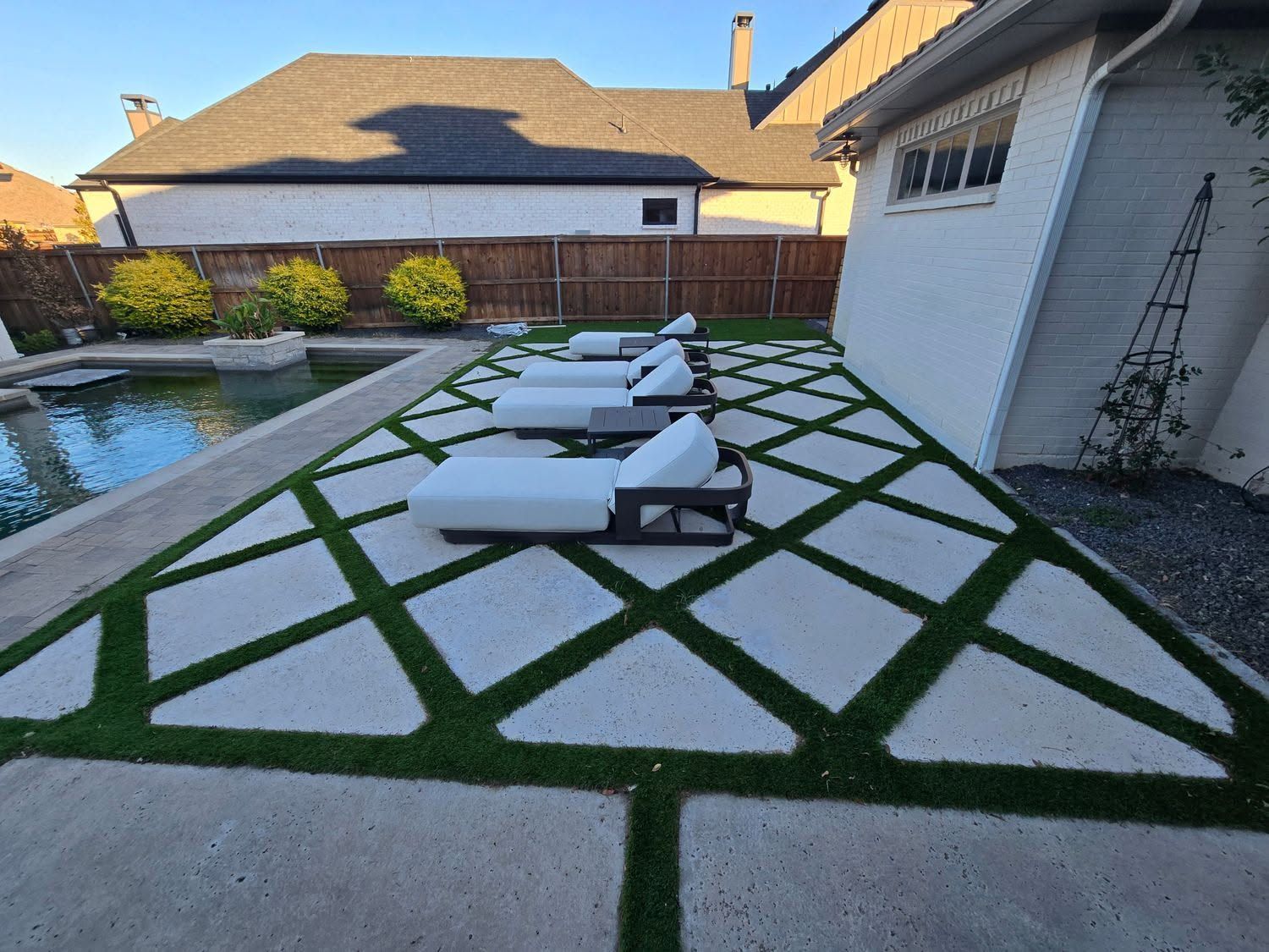Backyard patio with white lounge chairs, green grass pavers, and a swimming pool beside a house