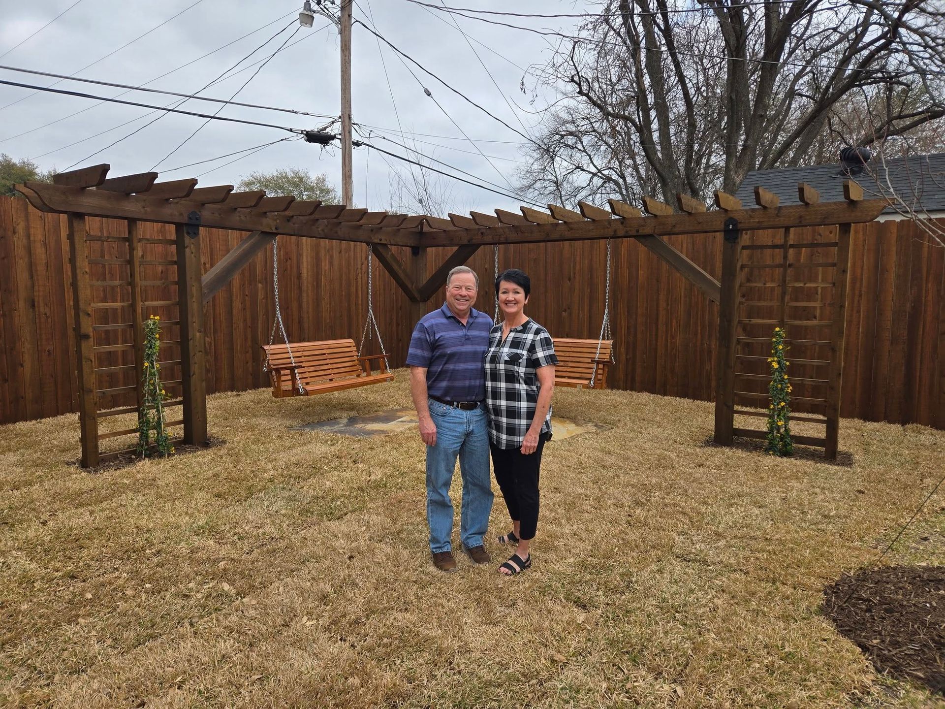 Two people standing together in a backyard with a wooden fence and leafless trees.