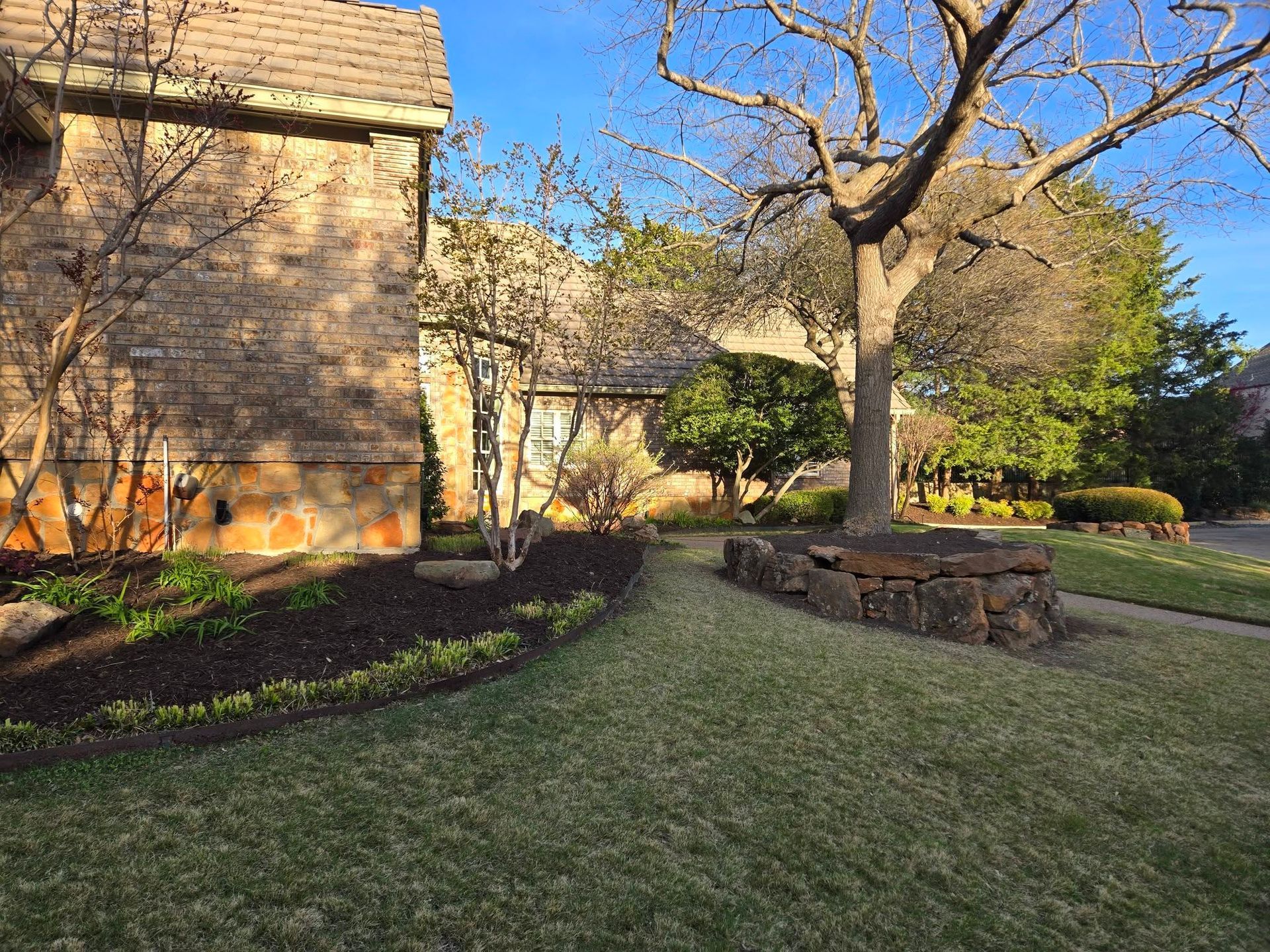 Stone house and lawn with leafless trees, a curved garden bed, and a brick retaining wall in warm sunlight